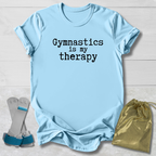 Light blue t-shirt with 'Gymnastics is my therapy' text, socks, and drawstring bag on a wooden surface.