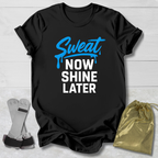 Black t-shirt with 'Sweat Now Shine Later' text, socks, and drawstring bag on a light wooden surface