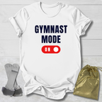 White t-shirt with 'GYMNAST MODE ON' text, socks, and drawstring bag on a light wooden surface