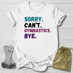 White t-shirt with colorful text 'Sorry. Can't. Gymnastics. Bye.' on a light wooden surface.