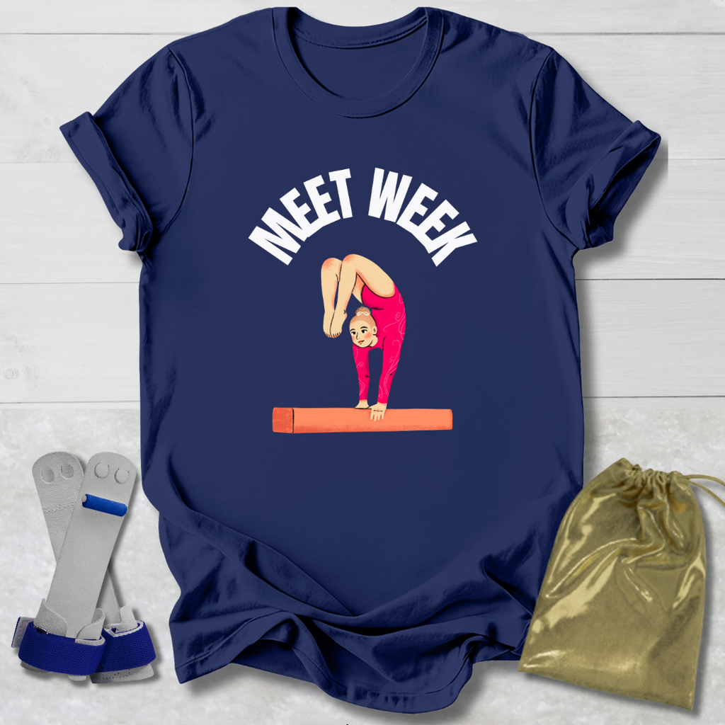 Navy blue t-shirt with a graphic of a gymnast on a balance beam and 'MEET WEEK' text, displayed with socks and a drawstring bag on a light wooden surface.