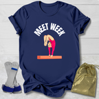 Navy blue t-shirt with a graphic of a gymnast on a balance beam and 'MEET WEEK' text, displayed with socks and a drawstring bag on a light wooden surface.