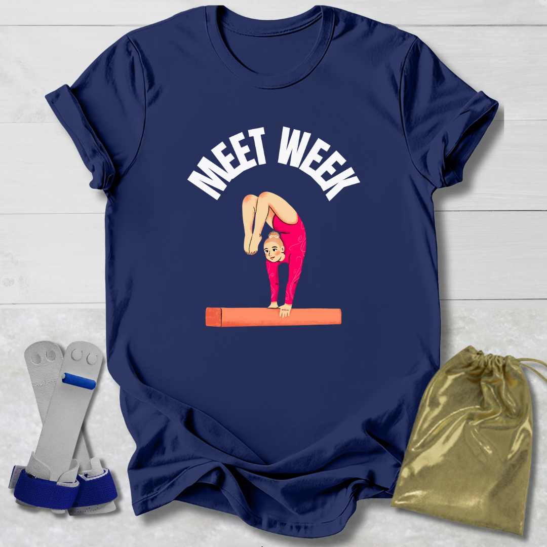 Navy blue t-shirt with a graphic of a gymnast on a balance beam and 'MEET WEEK' text, displayed with socks and a drawstring bag on a light wooden surface.