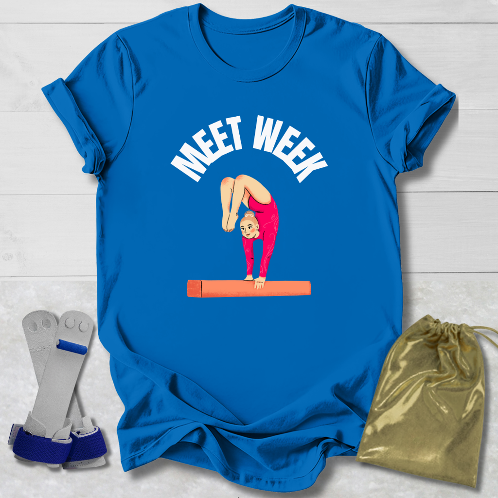 Blue t-shirt with a graphic of a gymnast and 'MEET WEEK' text, placed on a light wooden surface with socks and a drawstring bag.