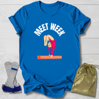 Blue t-shirt with a graphic of a gymnast and 'MEET WEEK' text, placed on a light wooden surface with socks and a drawstring bag.