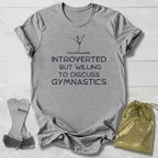 Gray t-shirt with 'Introverted but willing to discuss gymnastics' text on a wooden surface with socks and a drawstring bag.