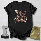 Black t-shirt with 'strong is the new pretty' text on a wooden surface with socks and a drawstring bag.