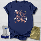 Navy blue t-shirt with 'Strong is the new pretty' text on a light wooden surface with socks and a drawstring bag.