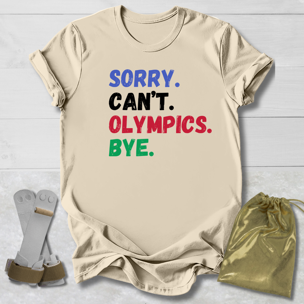 Beige t-shirt with colorful text 'SORRY. CAN'T. OLYMPICS. BYE.' on a light wooden surface.