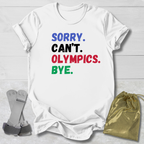 White t-shirt with colorful text 'SORRY. CAN'T. OLYMPICS. BYE.' on a light wooden surface.