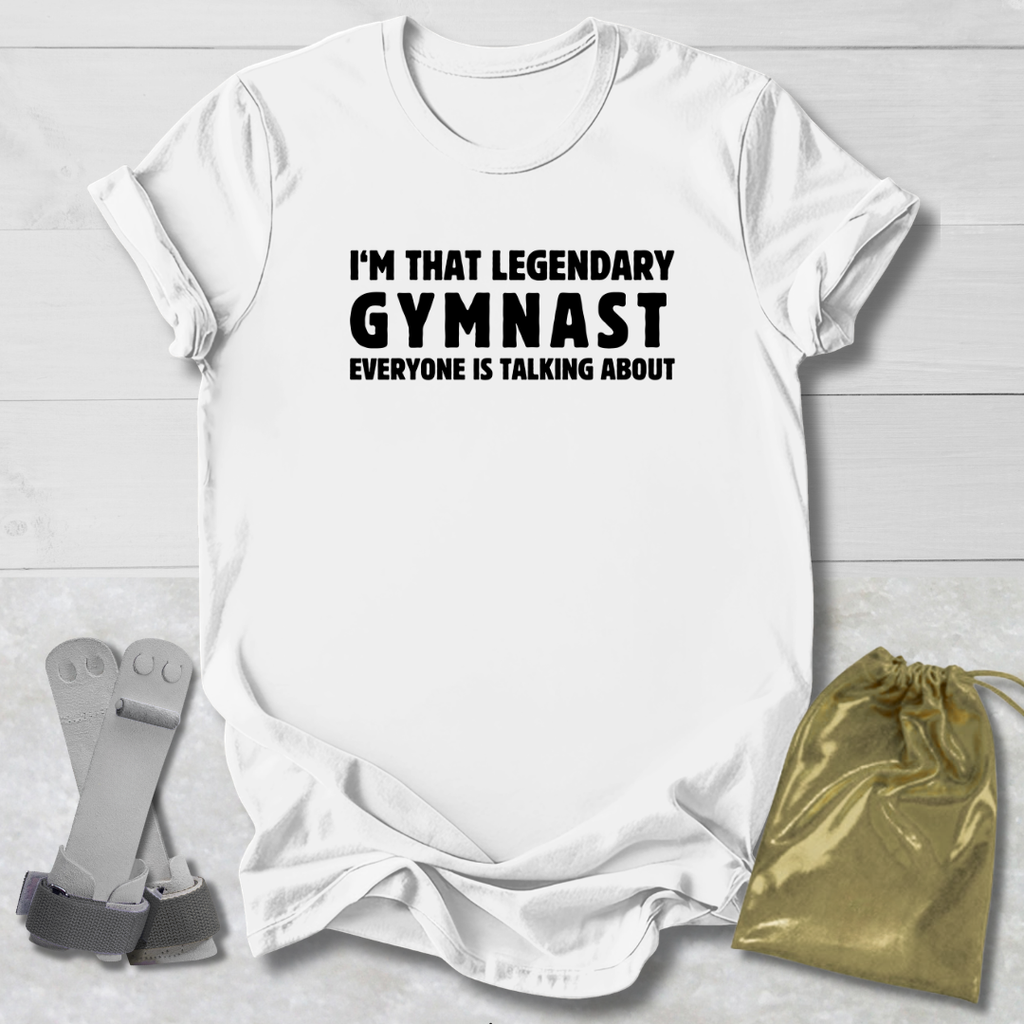 White t-shirt with text 'I'm that legendary gymnast everyone is talking about' on a light wooden surface, with socks and a drawstring bag.