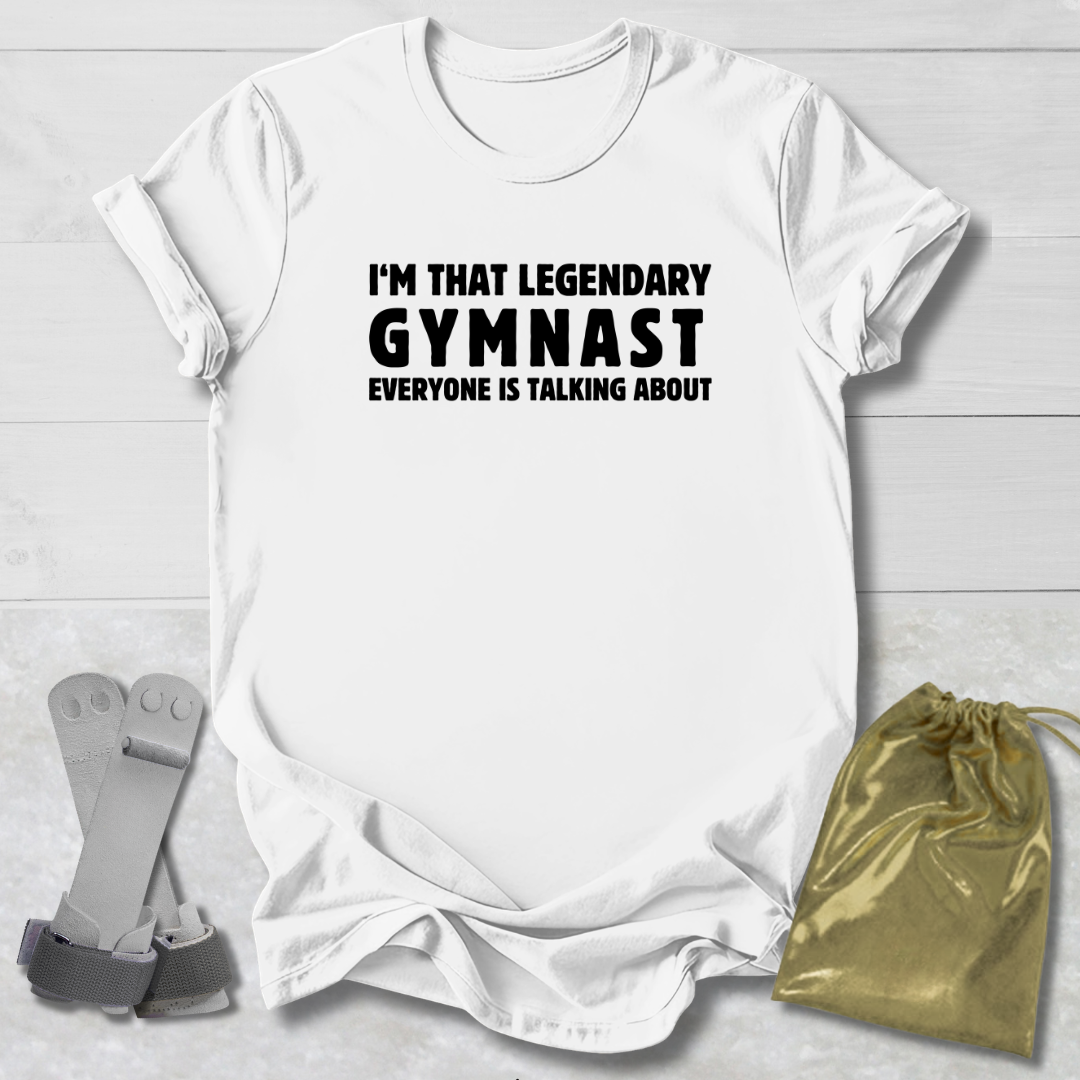 White t-shirt with text 'I'm that legendary gymnast everyone is talking about' on a light wooden surface, with socks and a drawstring bag.