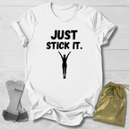 White gymnastics gymnast t-shirt with 'JUST STICK IT.' text and silhouette graphic on a light wooden surface.