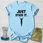 Light blue gymnastics gymnast t-shirt with 'JUST STICK IT.' text and silhouette, socks, and drawstring bag on a light wooden surface.
