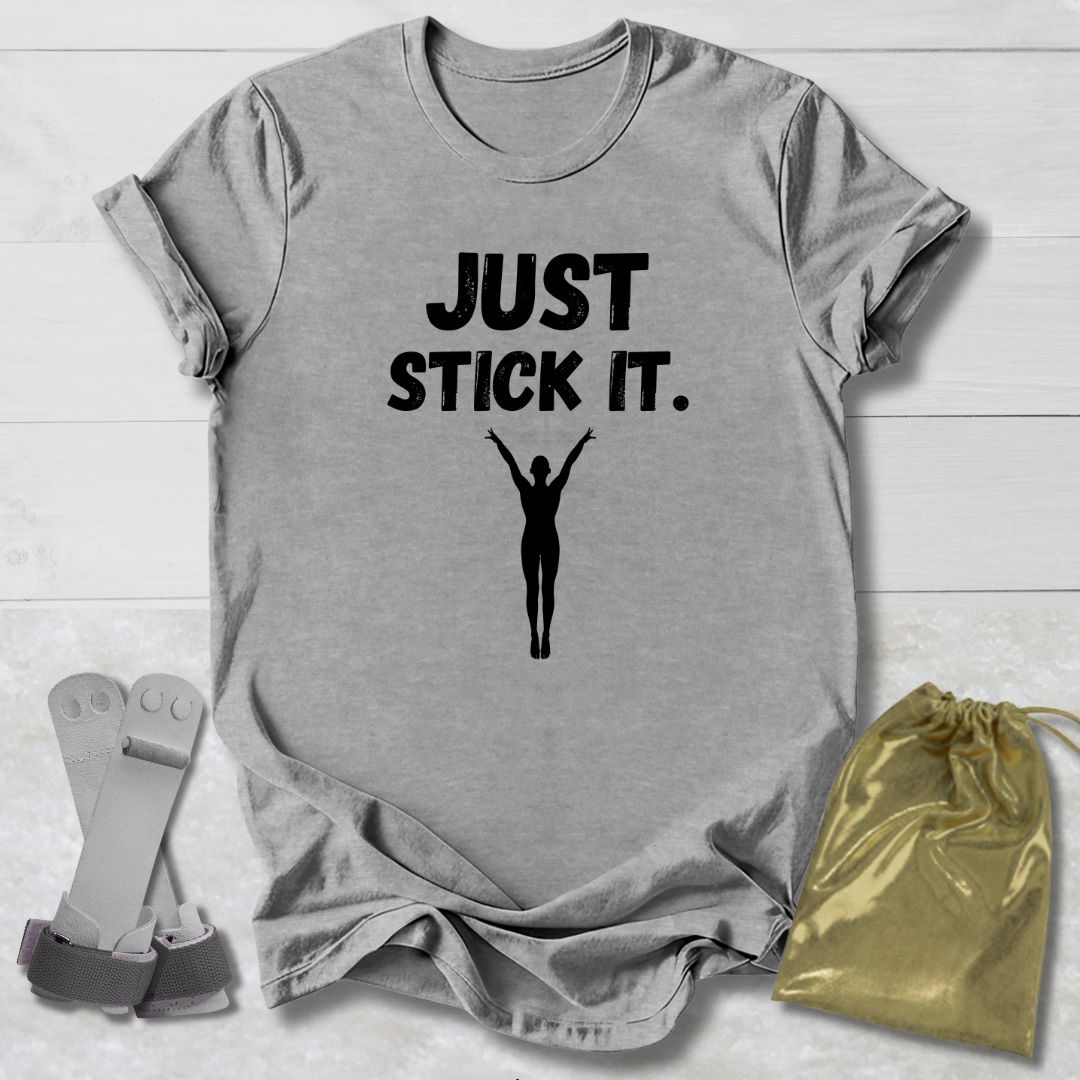 Gray gymnastics gymnast t-shirt with 'JUST STICK IT.' text and silhouette graphic on a wooden surface with socks and a drawstring bag.