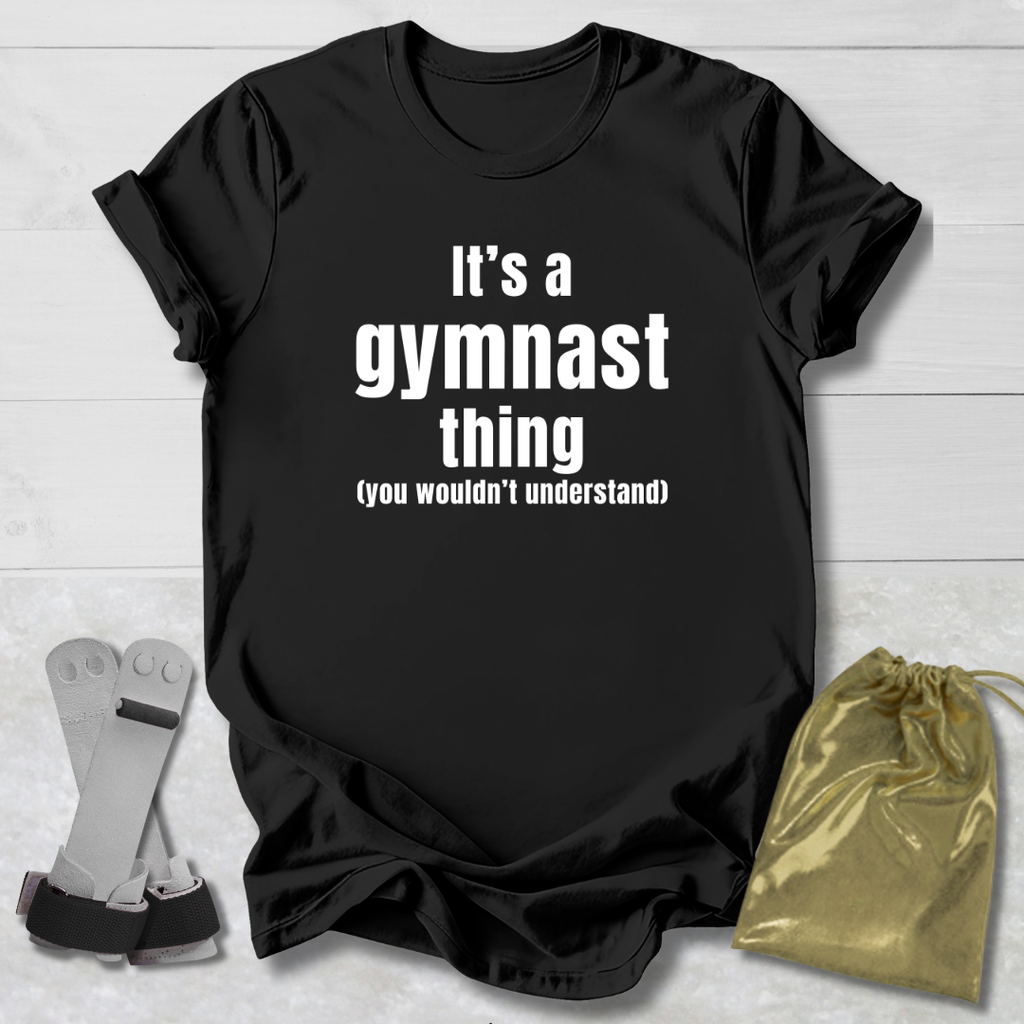 Black t-shirt with 'It's a gymnast thing (you wouldn't understand)' text on a light wooden surface with socks and a drawstring bag.