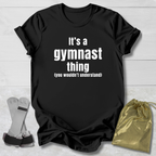 Black t-shirt with 'It's a gymnast thing (you wouldn't understand)' text on a light wooden surface with socks and a drawstring bag.