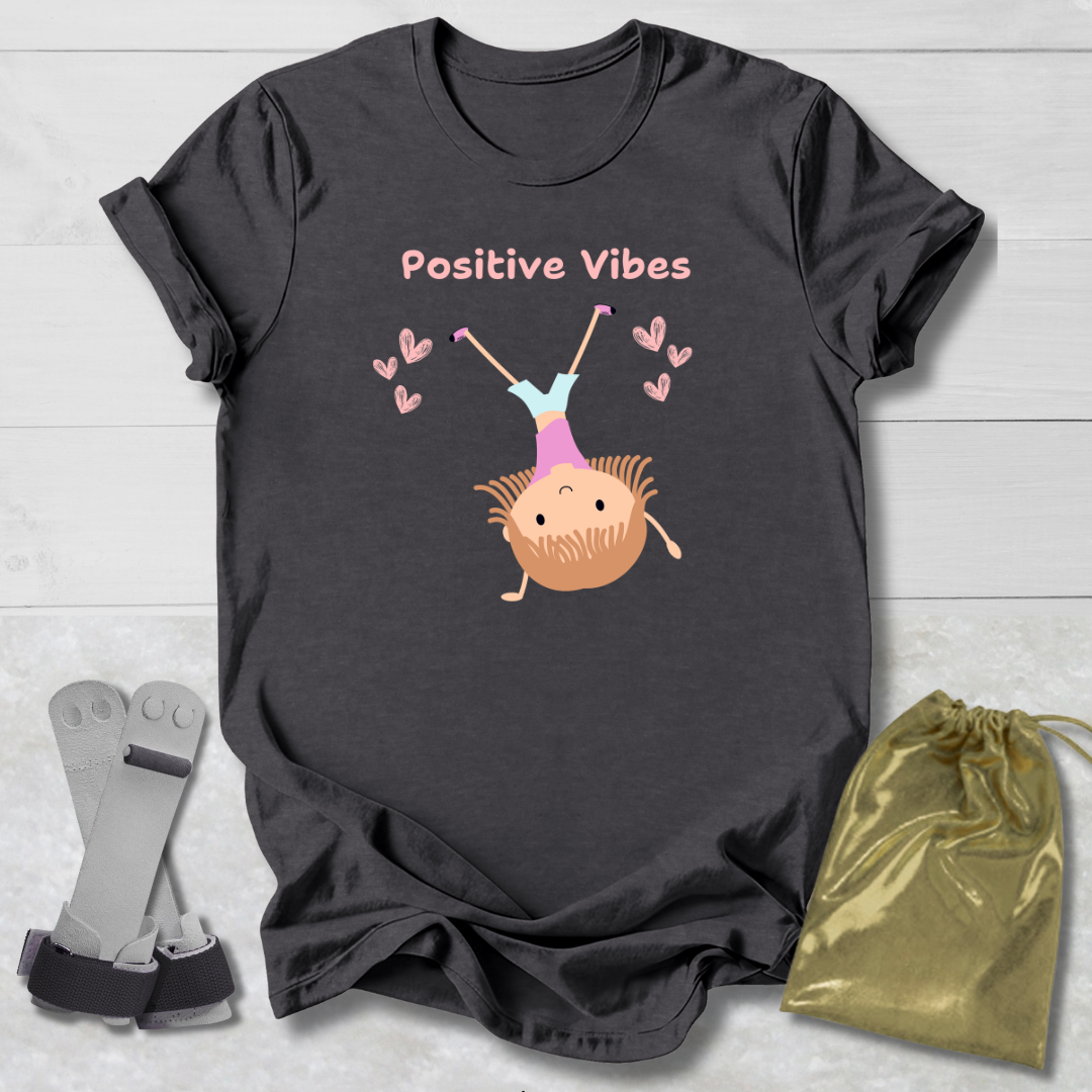 Dark gray t-shirt with 'Positive Vibes' and cartoon character on a light wooden surface with socks and a drawstring bag.