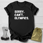 Black t-shirt with 'Sorry. Can't. Olympics.' text on a light wooden surface with socks and a drawstring bag.