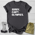 Black t-shirt with 'Sorry. Can't. Olympics.' text on a light wooden surface with socks and a drawstring bag.