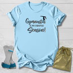 Light blue t-shirt with 'Gymnastics is my favorite season!' text, socks, and drawstring bag on a wooden surface.