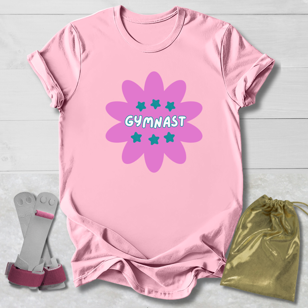 Pink t-shirt with purple flower design and 'GYMNAST' text, paired with gray socks and a gold drawstring bag on a light wooden surface.