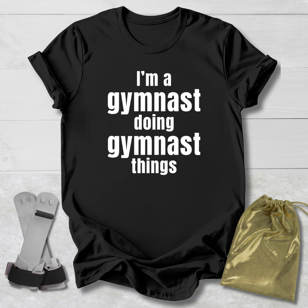 Black t-shirt with 'I'm a gymnast doing gymnast things' text, socks, and drawstring bag on a light wooden surface.