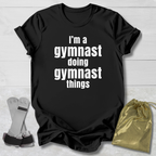 Black t-shirt with 'I'm a gymnast doing gymnast things' text, socks, and drawstring bag on a light wooden surface.