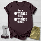 Maroon t-shirt with 'I'm a gymnast doing gymnast things' text, socks, and drawstring bag on a light wooden surface.