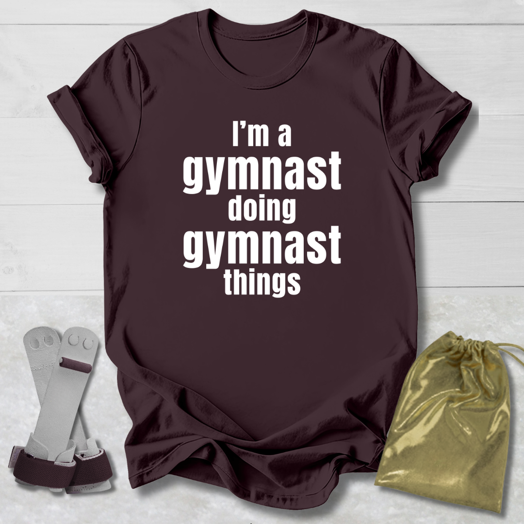 Maroon t-shirt with 'I'm a gymnast doing gymnast things' text, socks, and drawstring bag on a light wooden surface.