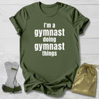 Green t-shirt with 'I'm a gymnast doing gymnast things' text, socks, and drawstring bag on a light wooden surface.