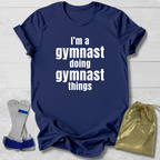 Navy blue t-shirt with 'I'm a gymnast doing gymnast things' text, socks, and drawstring bag on a light wooden surface.