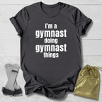 Gray t-shirt with 'I'm a gymnast doing gymnast things' text, socks, and drawstring bag on a light wooden surface.