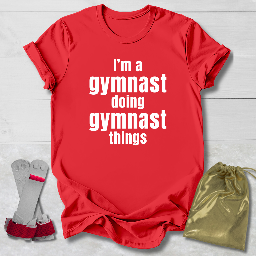 Red t-shirt with 'I'm a gymnast doing gymnast things' text, socks, and drawstring bag on a light wooden surface.