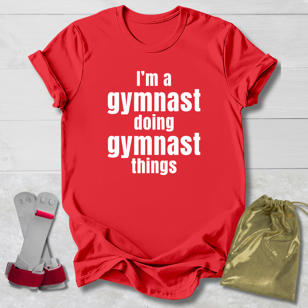 Red t-shirt with 'I'm a gymnast doing gymnast things' text, socks, and drawstring bag on a light wooden surface.