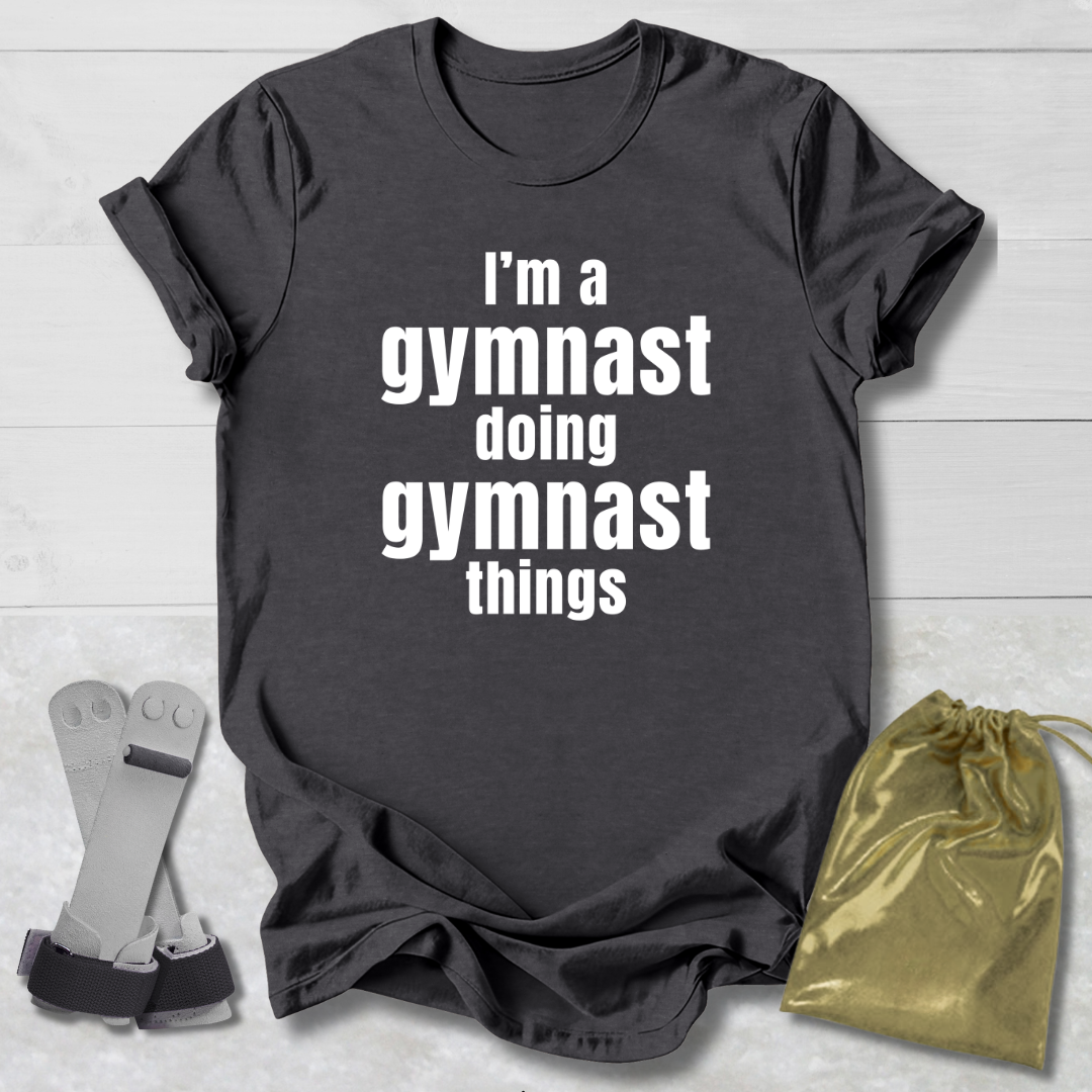 Gray t-shirt with 'I'm a gymnast doing gymnast things' text, socks, and drawstring bag on a light wooden surface.
