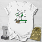 White t-shirt with a cartoon design of a rabbit on a bench with a basket, socks, and a drawstring bag on a light wooden surface.