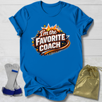 Blue t-shirt with 'I'm the Favorite Coach' design on a light wooden surface, with socks and a drawstring bag in the foreground.