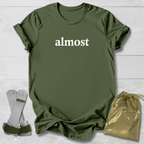 Green t-shirt with 'ALMOST.' text, socks, and drawstring bag on a light wooden surface