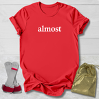 Red t-shirt with 'ALMOST.' text, socks, and drawstring bag on a light wooden surface