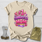 Beige t-shirt with '#1 Gymnastics Mom' text and graphics on a light wooden surface.