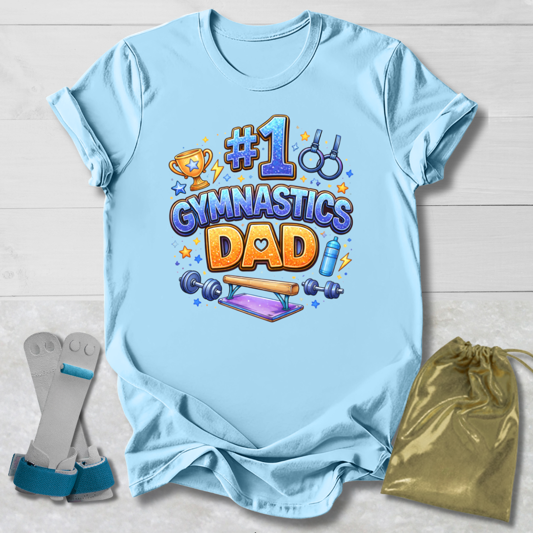 Light blue t-shirt with '#1 Gymnastics Dad' design on a wooden surface with socks and a drawstring bag.