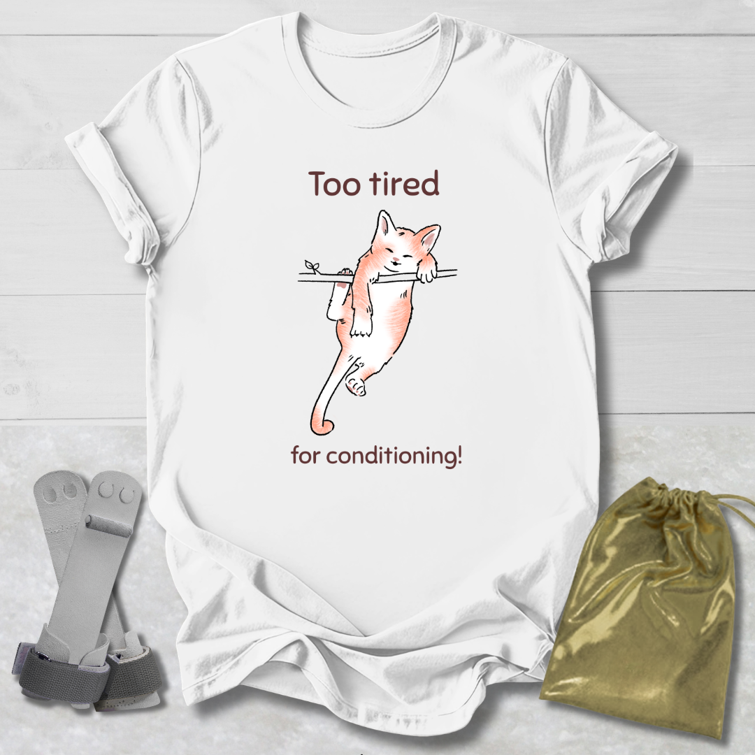 White t-shirt with a cat illustration and a cat that's too tired for gymnastics conditioning text, paired with socks and a drawstring bag on a light wooden surface.