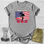 Gray t-shirt with Gymnast American flag and 'Made in the USA' text on a wooden surface, with socks and a drawstring bag in the foreground.