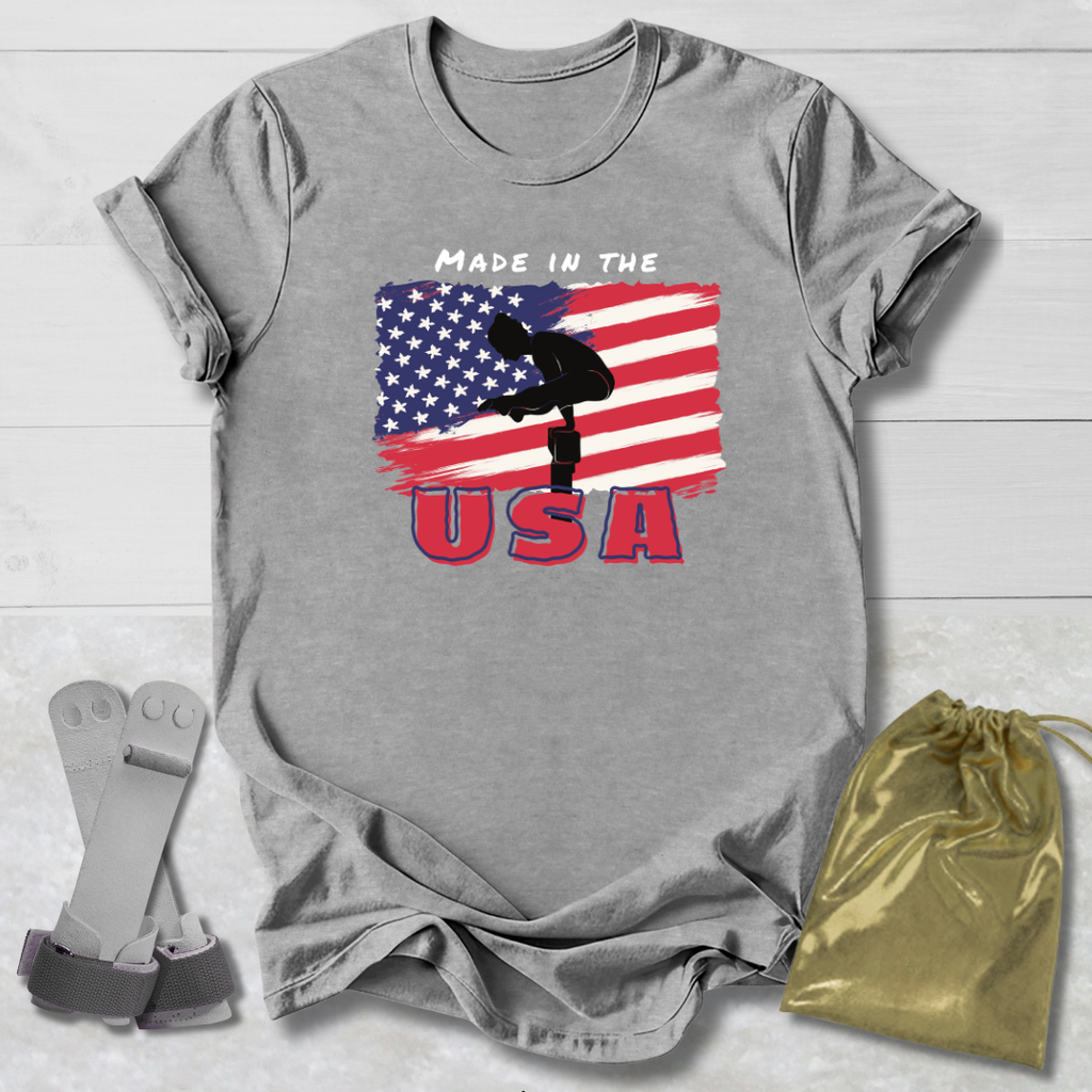 Gray t-shirt with Gymnast American flag and 'Made in the USA' text on a wooden surface, with socks and a drawstring bag in the foreground.