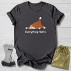 Dark gray t-shirt with a cartoon dog and 'Everything Hurts' text, placed on a light wooden surface with socks and a drawstring bag.