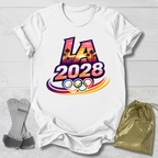 White t-shirt with 'LA 2028' Olympic design on a light wooden surface with socks and a drawstring bag.