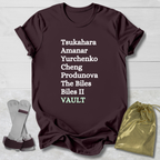 Maroon t-shirt with gymnast vault skills text on a light wooden surface with socks and a drawstring bag.