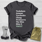 Dark gray t-shirt with white gymnast vault skills text on a light wooden surface with socks and a drawstring bag.