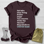 Brown t-shirt with gymnast uneven bars skills text on a light wooden surface with socks and a drawstring bag.
